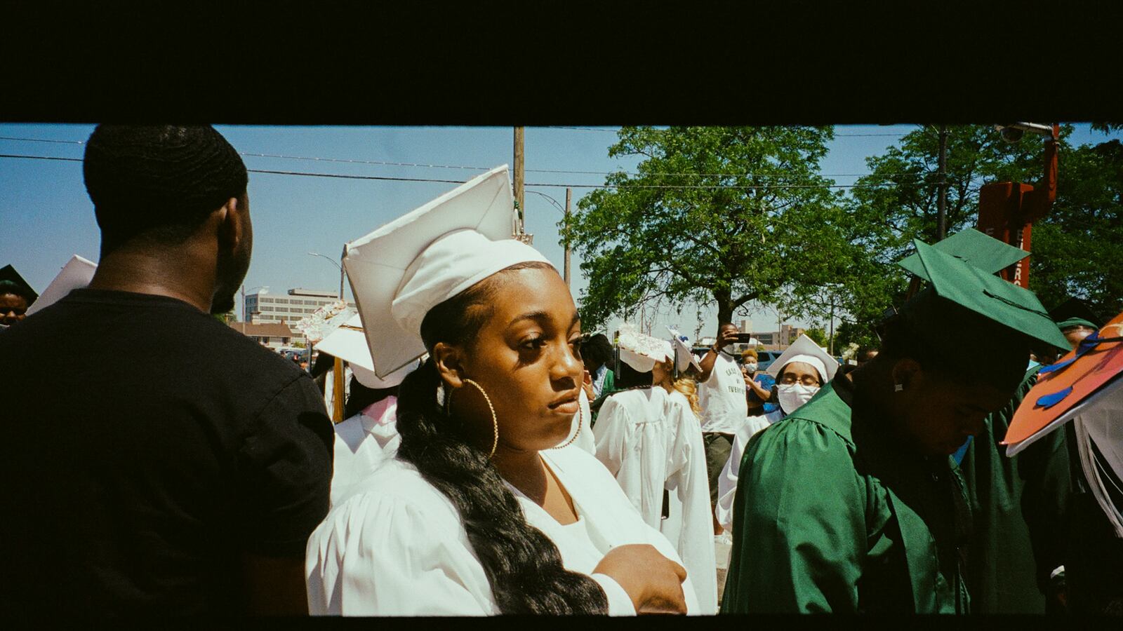 A young woman wearing a white graduation gown and cap looks contemplative as she’s surrounded by other teenagers.