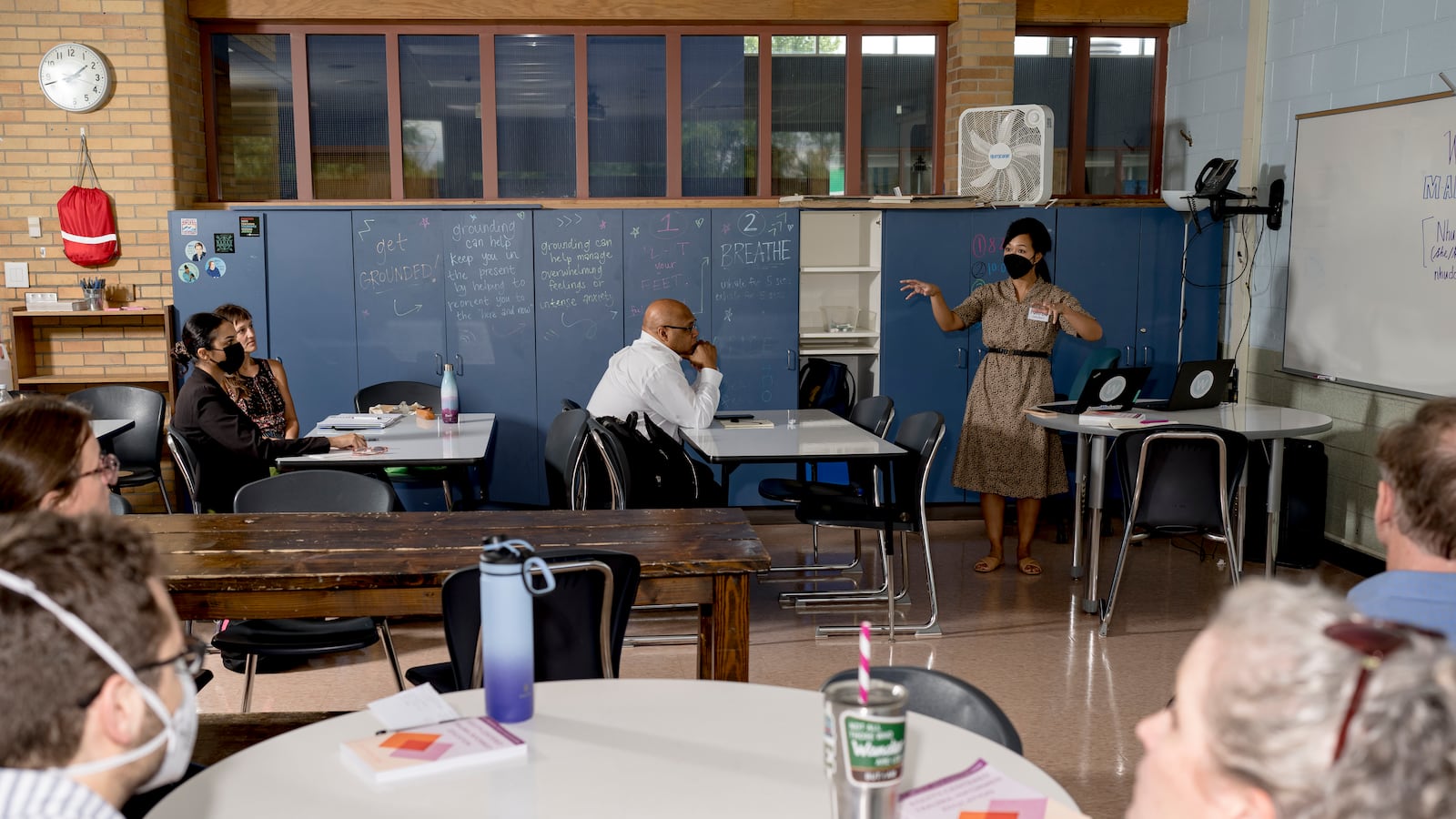 A principal speaks in front of a half dozen adults seated at round tables.