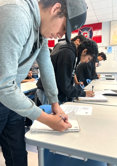A male student in a gray sweatshirt and cap writes on a paper on a table with other students.