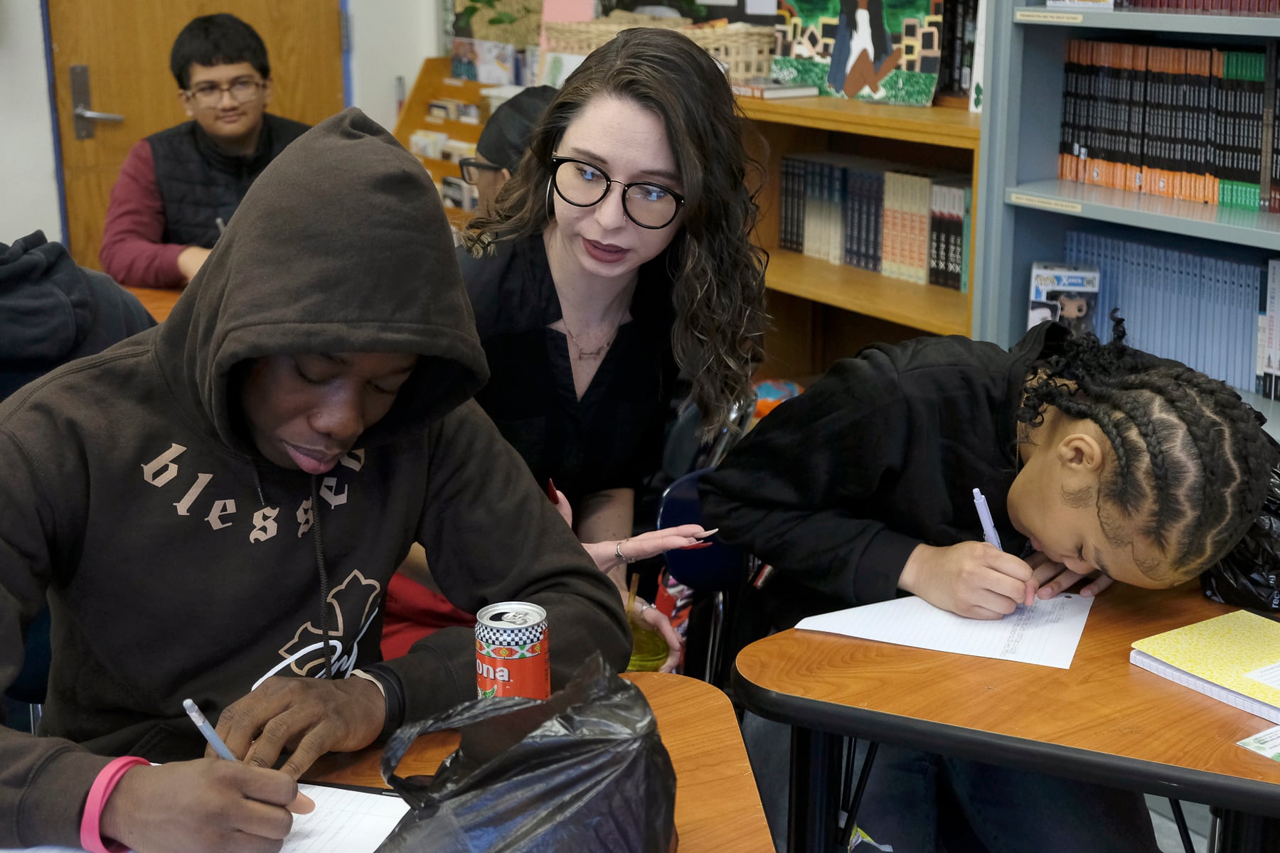 A white woman teacher leans between two Black high school students who are working at their wooden desks in a classroom.