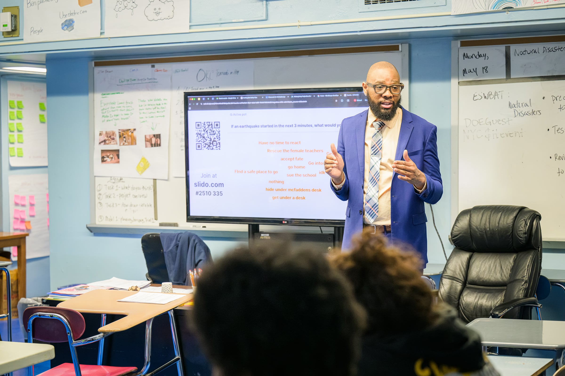 A Black man in a suit stands at the front of a classroom next to a large screen.