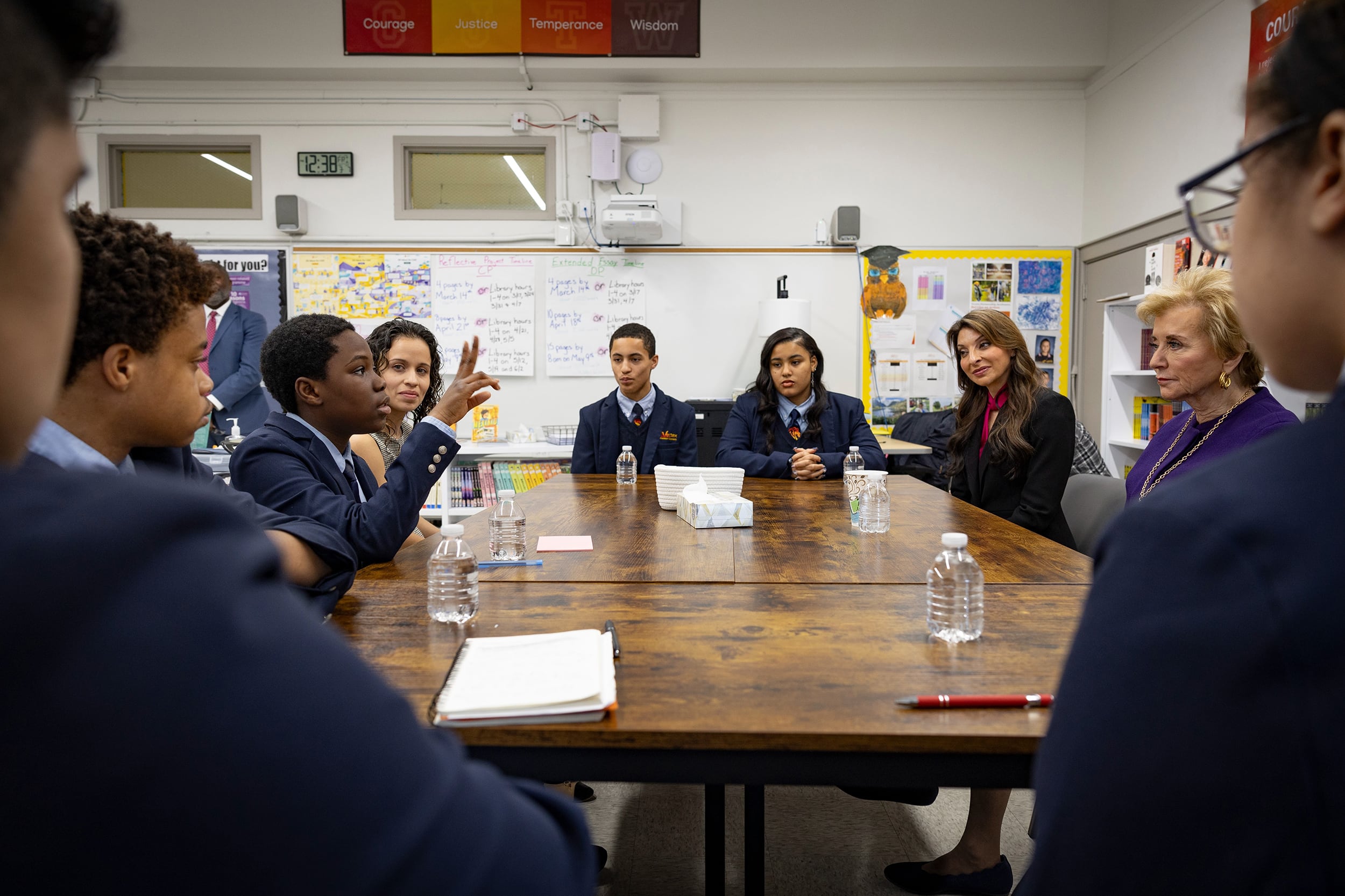 A large group of older students all wearing the same dark blue uniform sit around a wooden table talking with two white women in suits.