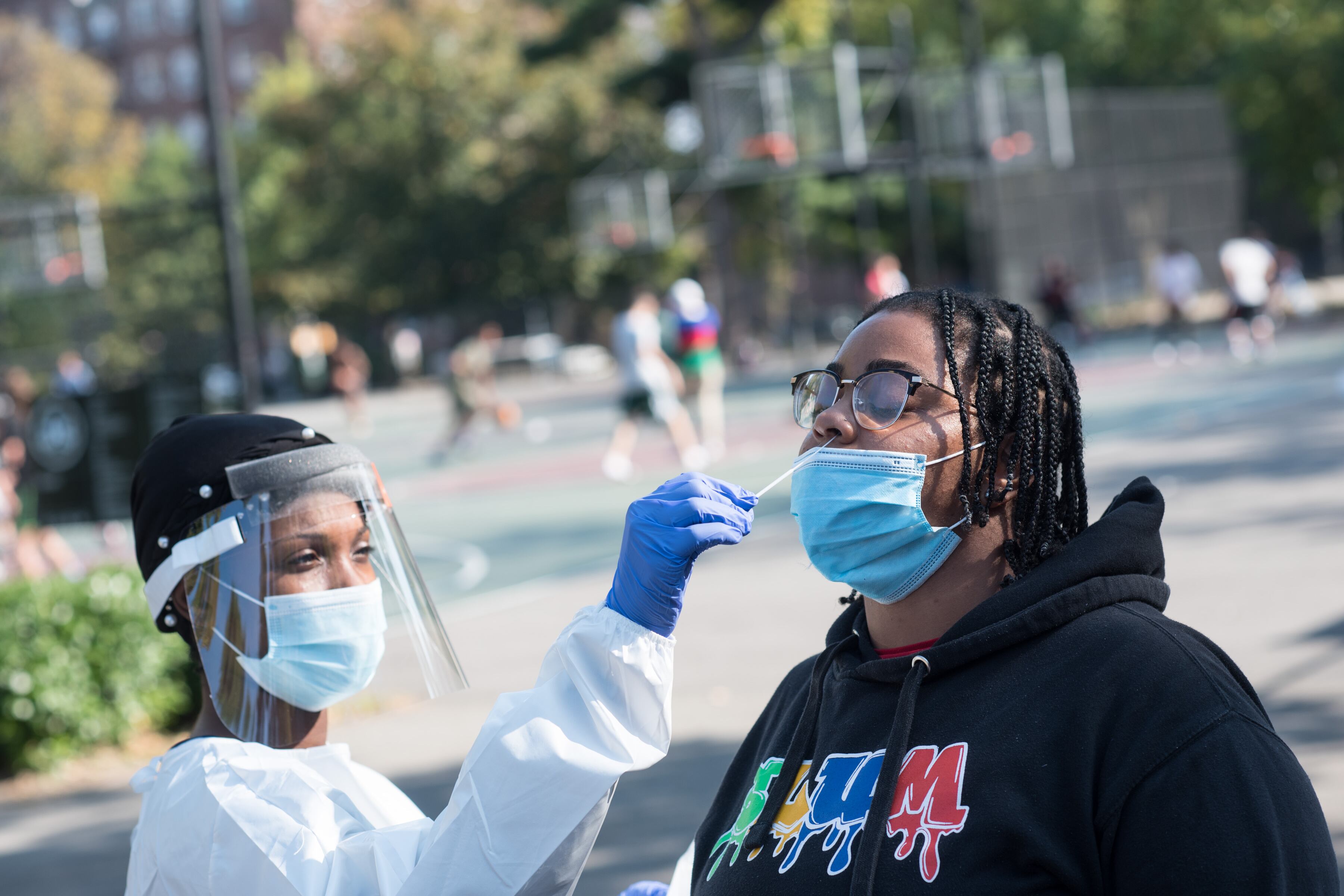 A woman with a face shield and masks swabs the nose of another masked woman outdoors.
