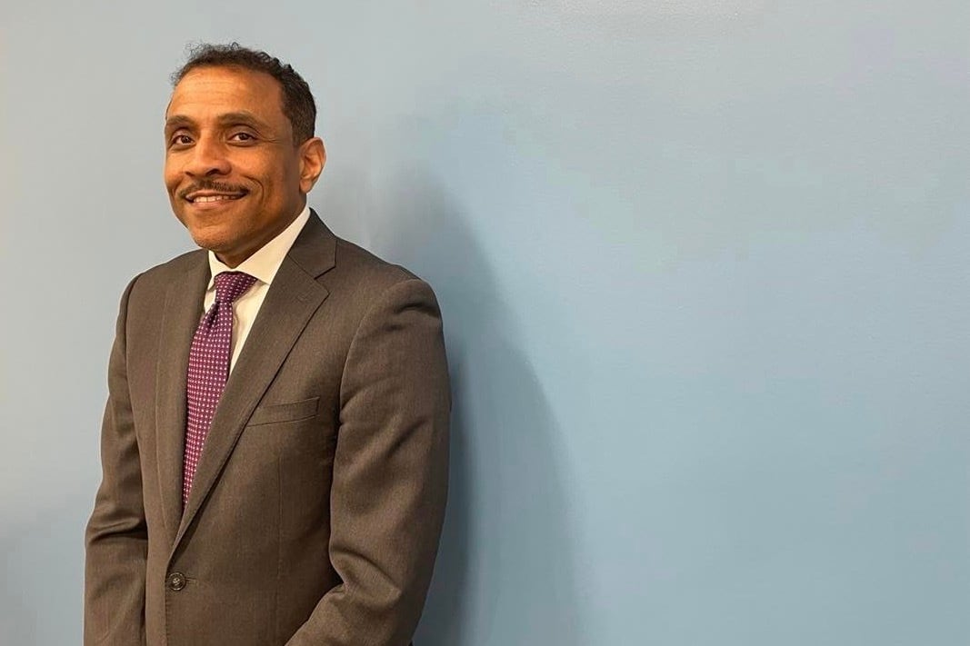 A man in a brown suit and a maroon tie stands in front of a pale blue background and smiles.