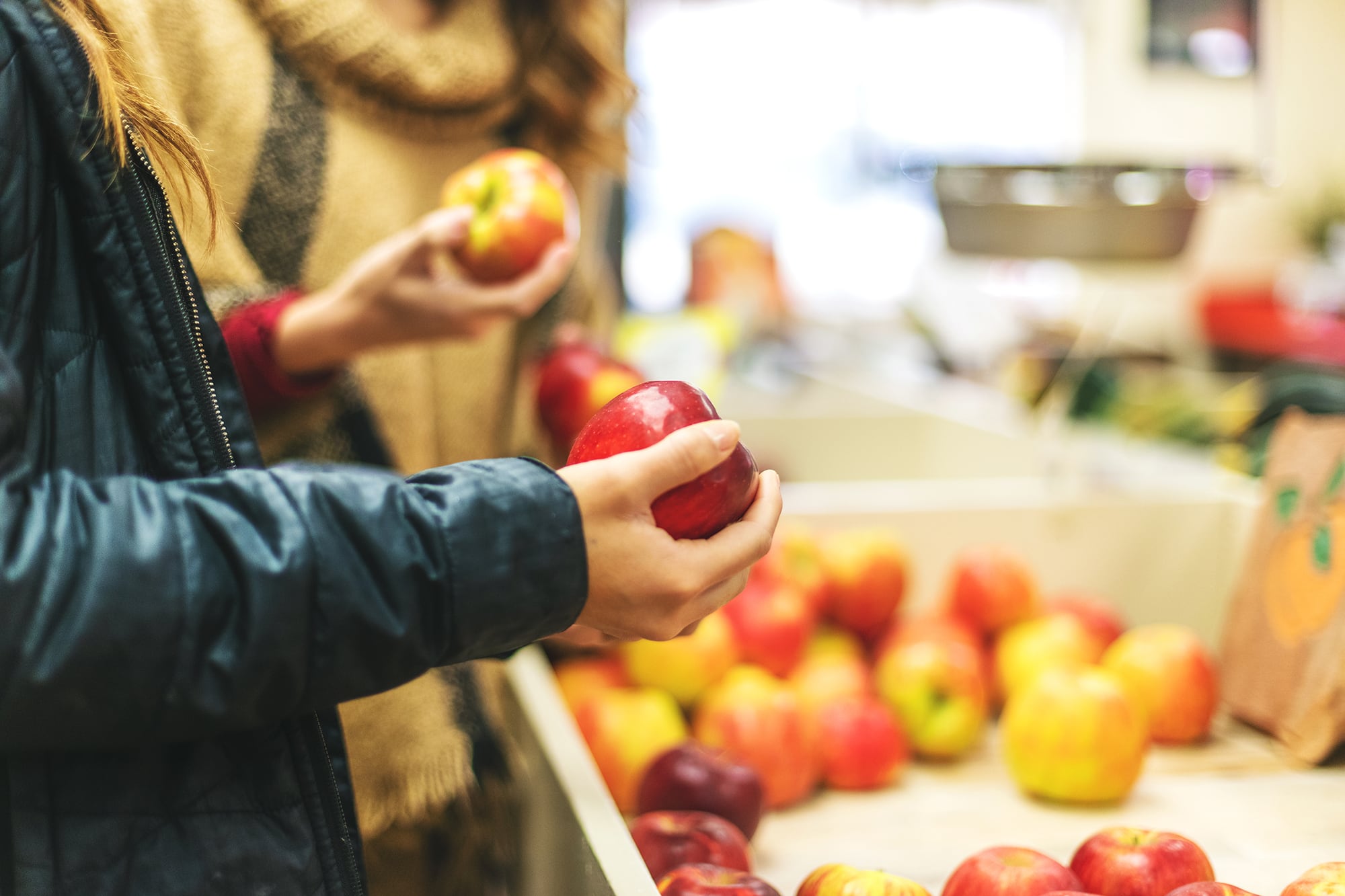 An adult arm with a black leather jacket holds a red apple with another adult holding a red and yellow apple in the background with apples in a container.