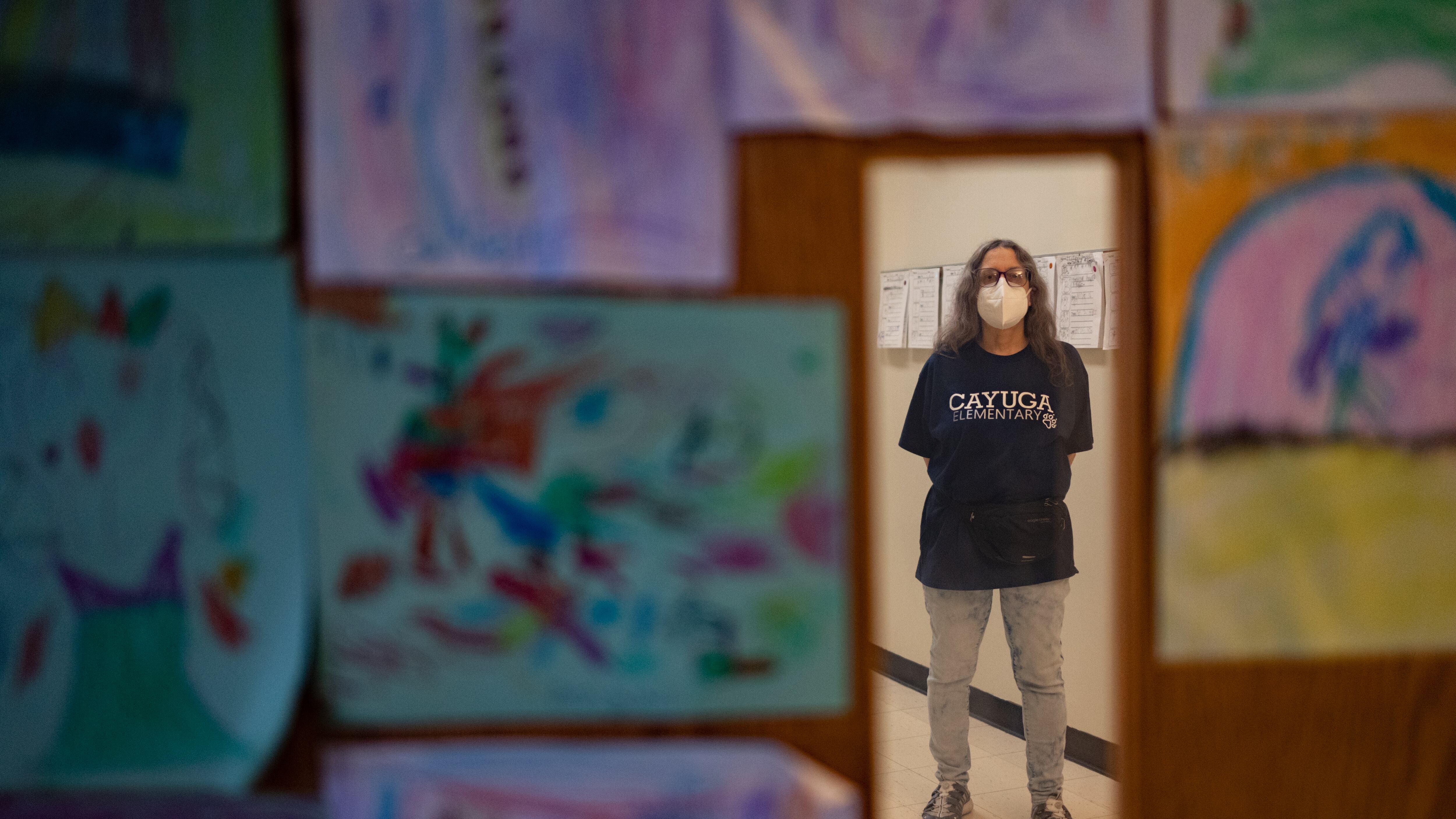An older woman, wearing a filtered mask and blue Cayuga Elementary School shirt, stands in the window of a classroom door. Student artwork adds color to the wooden door she is standing behind.