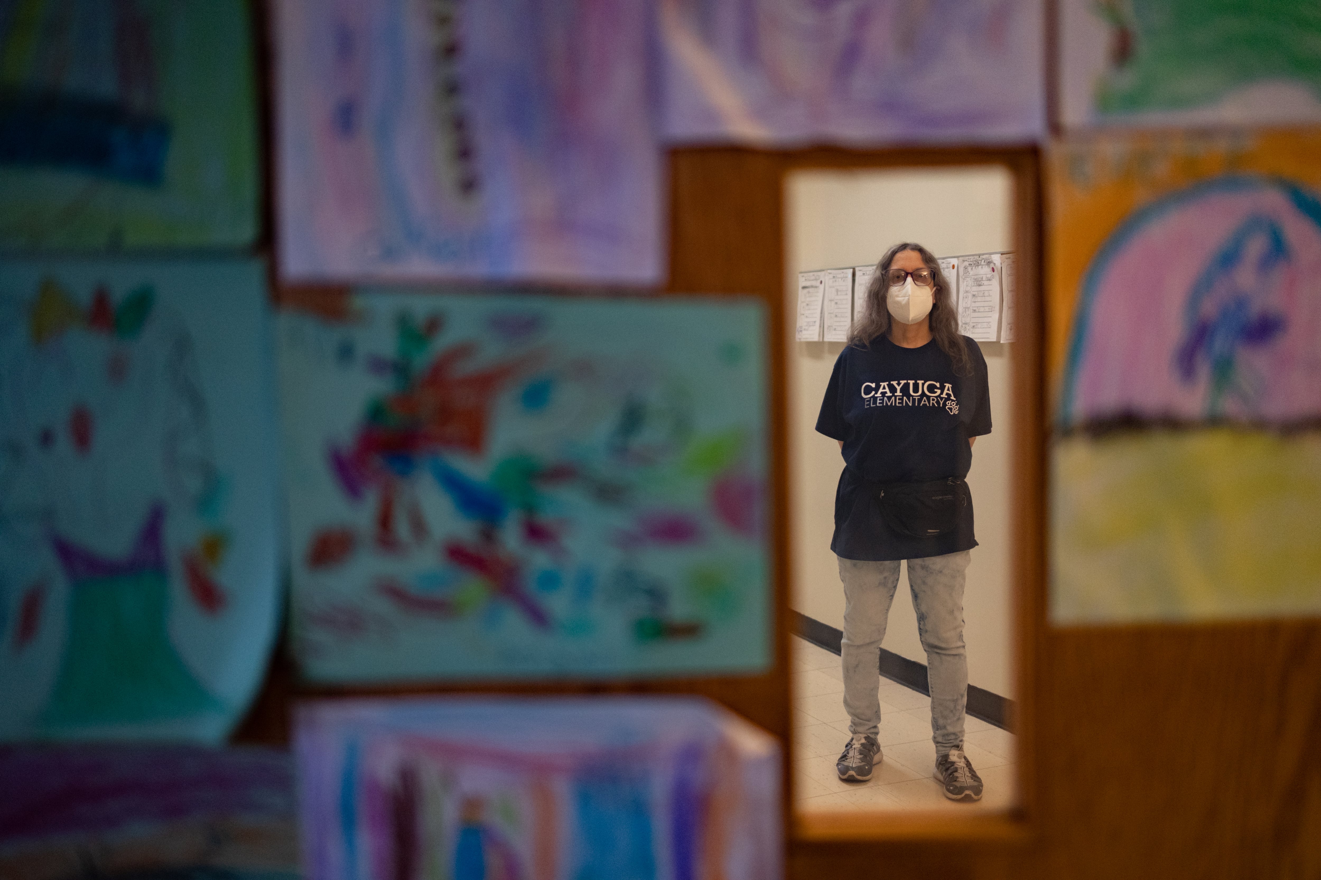 An older woman, wearing a filtered mask and blue Cayuga Elementary School shirt, stands in the window of a classroom door. Student artwork adds color to the wooden door she is standing behind.