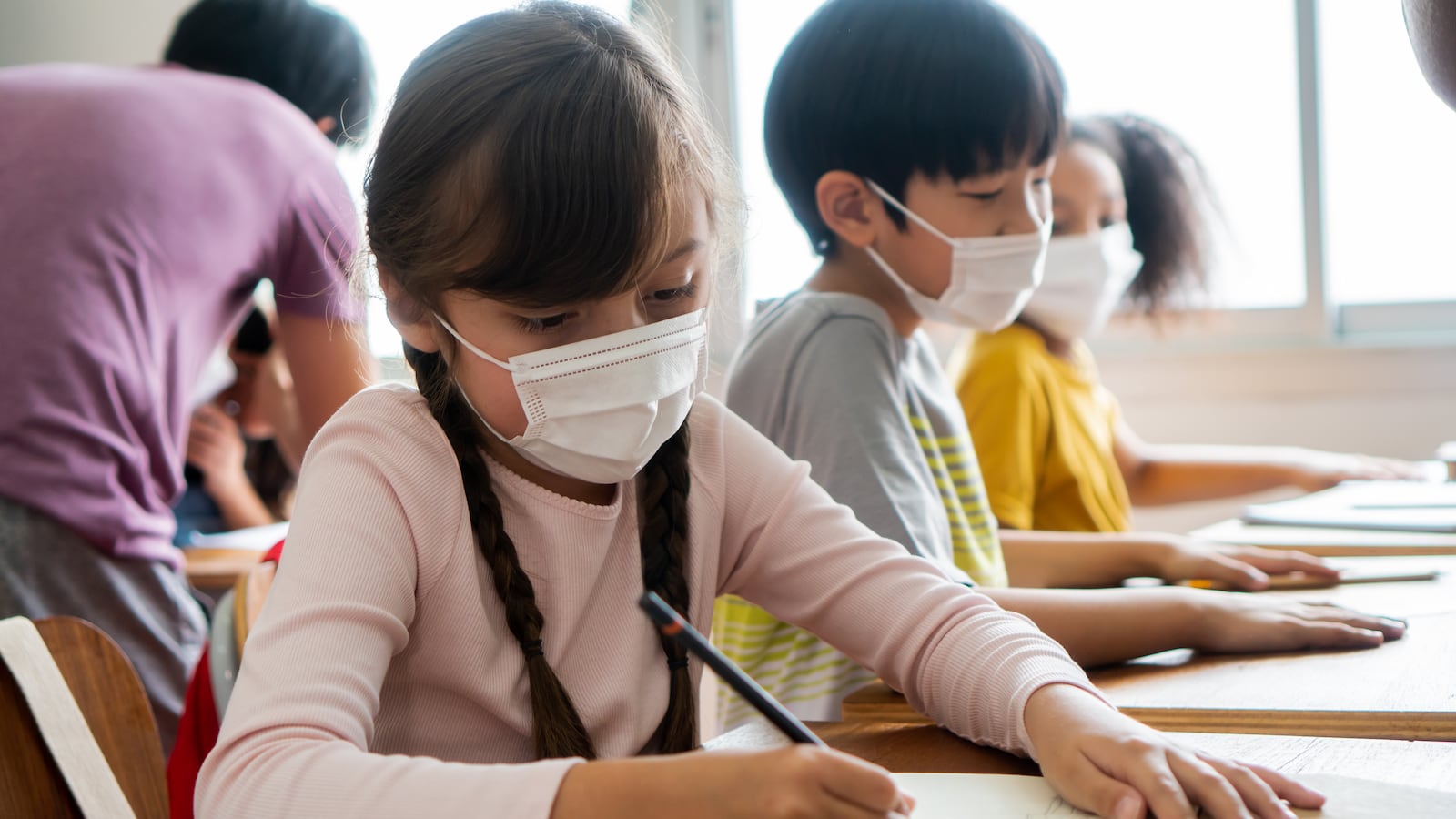 Multiethnic group of school children wearing face coverings sitting at school desks, writing.