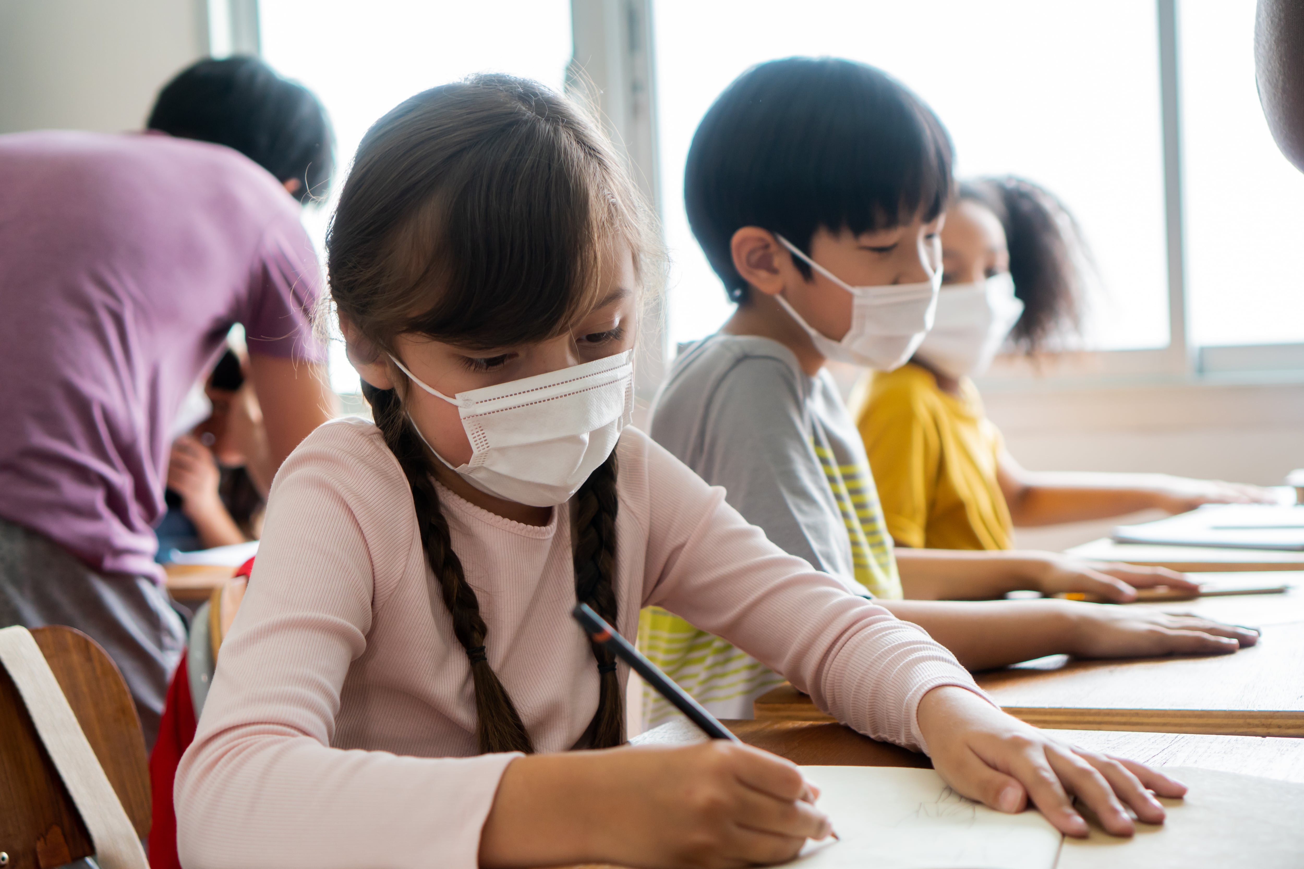 Multiethnic group of school children wearing face coverings sitting at school desks, writing.