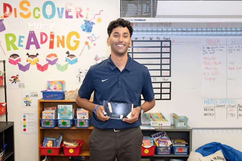 A man in a blue shirt holds an object in front of a bulletin board.