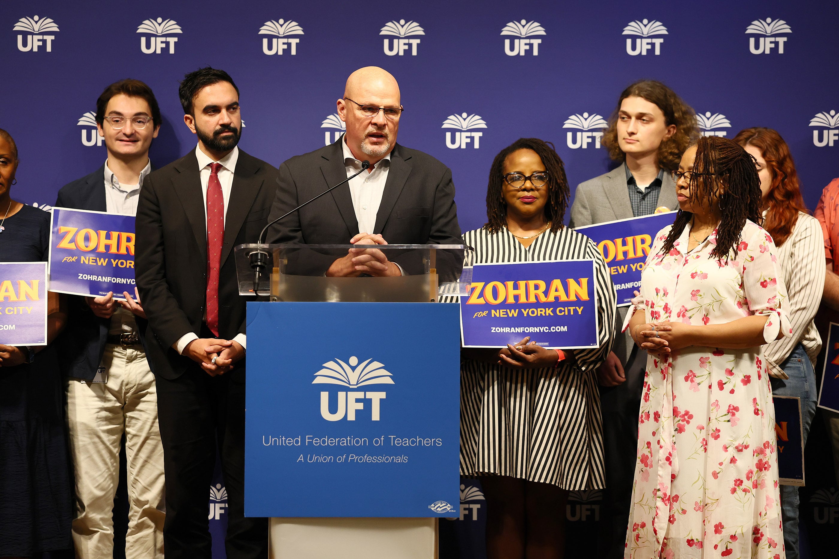 A white man in a suit speaks from behind a podium with a group of people standing behind him all on a stage.