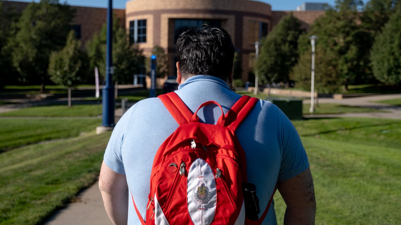 A student wearing a blue shirt and red backpack walks down a sidewalk on a college campus.