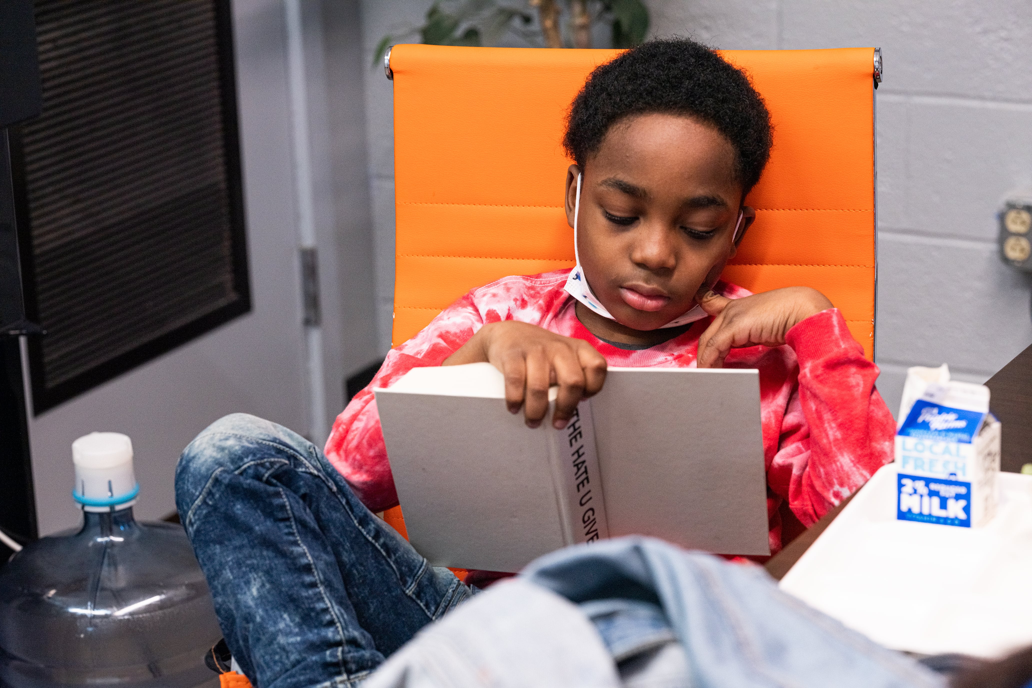 A student sits in a chair with one leg propped up and a carton of milk nearby, reading a hardcover copy of “The Hate U Give” with its book jacket taken off.