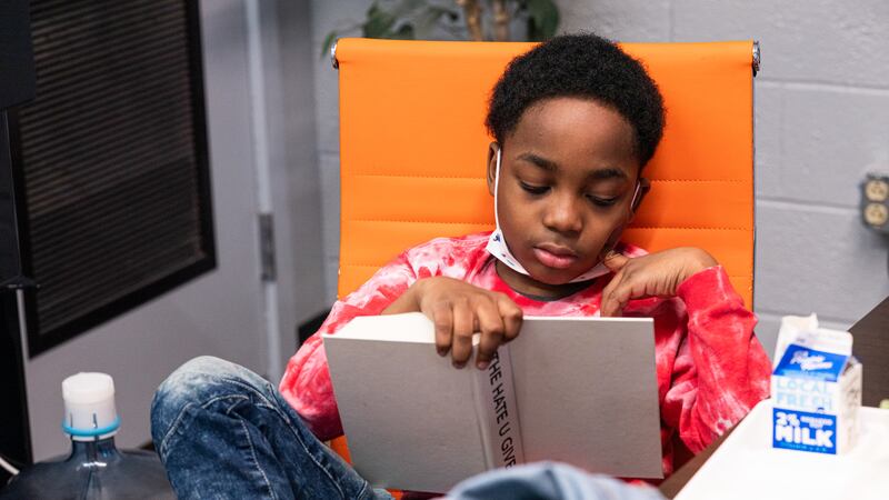 A student sits in a chair with one leg propped up and a carton of milk nearby, reading a hardcover copy of “The Hate U Give” with its book jacket taken off.