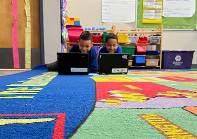 Two students lay on a colorful rug working on computers with a toy shelf in the background.