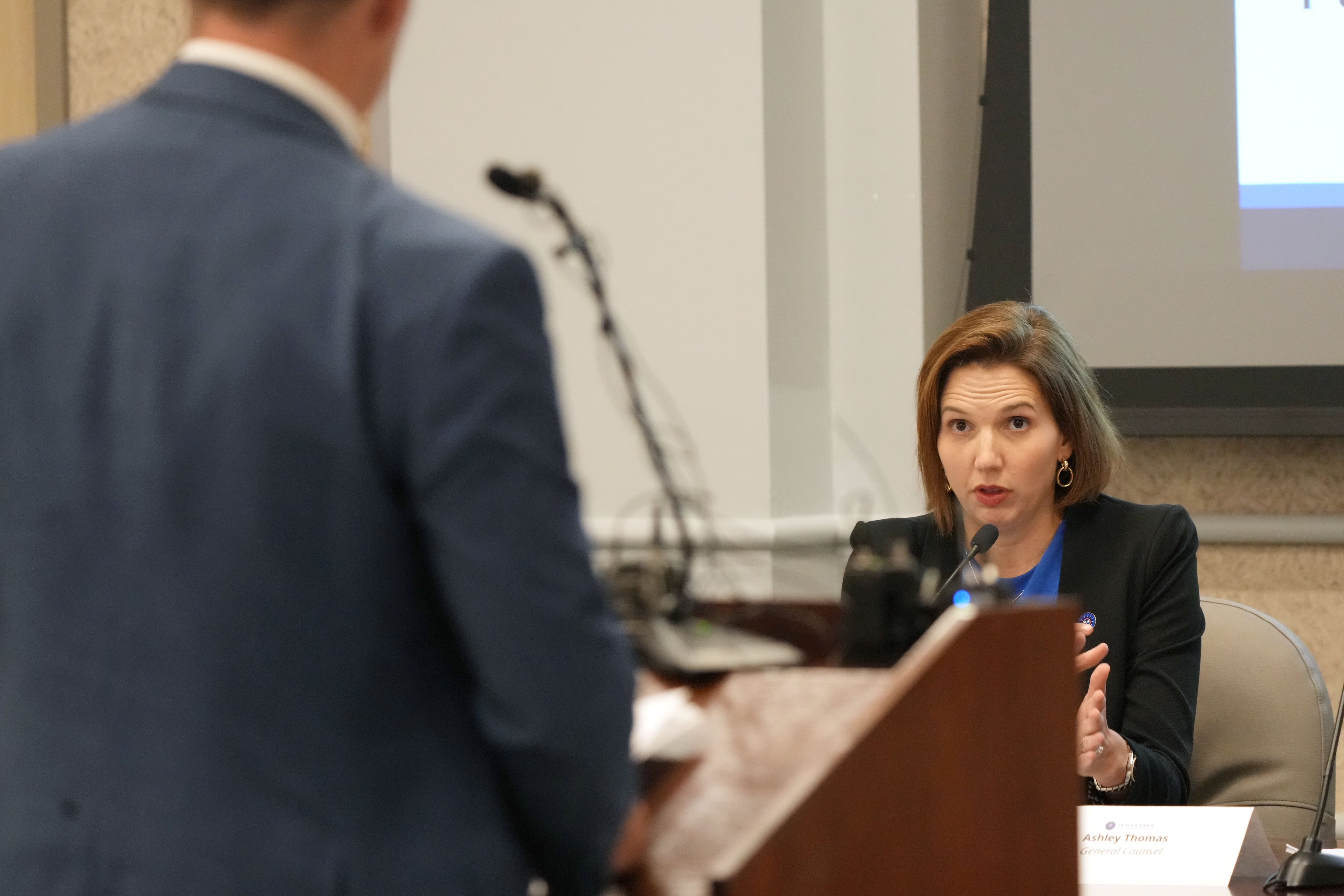 A man speaks from a podium while a woman sitting at a desk asks questions.