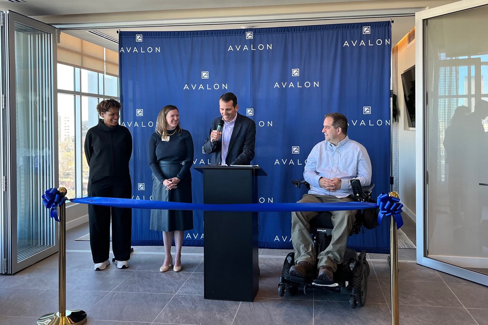 Three people stand and one person sits in a chair behind a podium and in front of a blue background during a ribbon cutting ceremony.