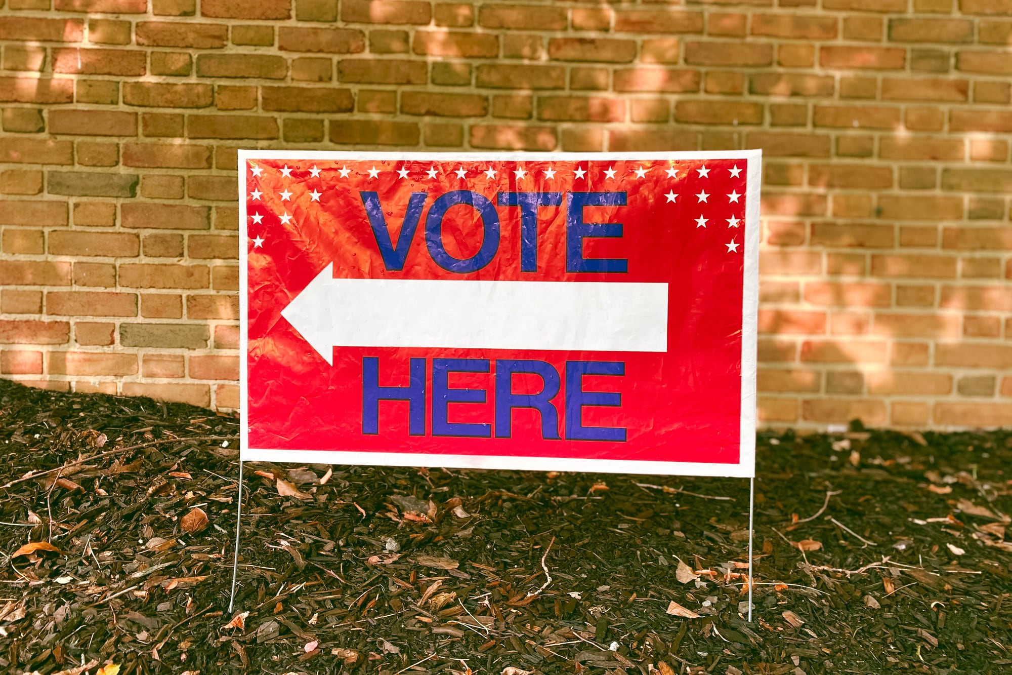 A red sign reads "Vote Here" with a tan brick wall in the background.
