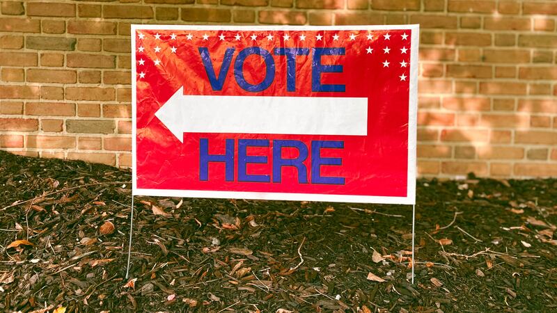 A red sign reads "Vote Here" with a tan brick wall in the background.