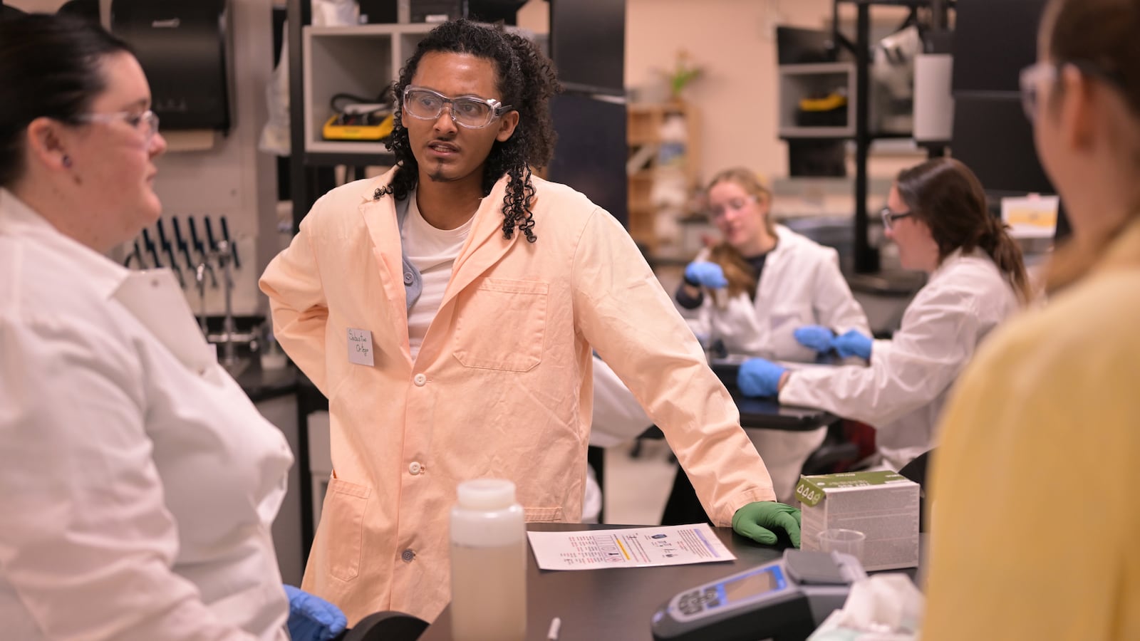 Students wearing lab coats and safety equipment work in a college laboratory.