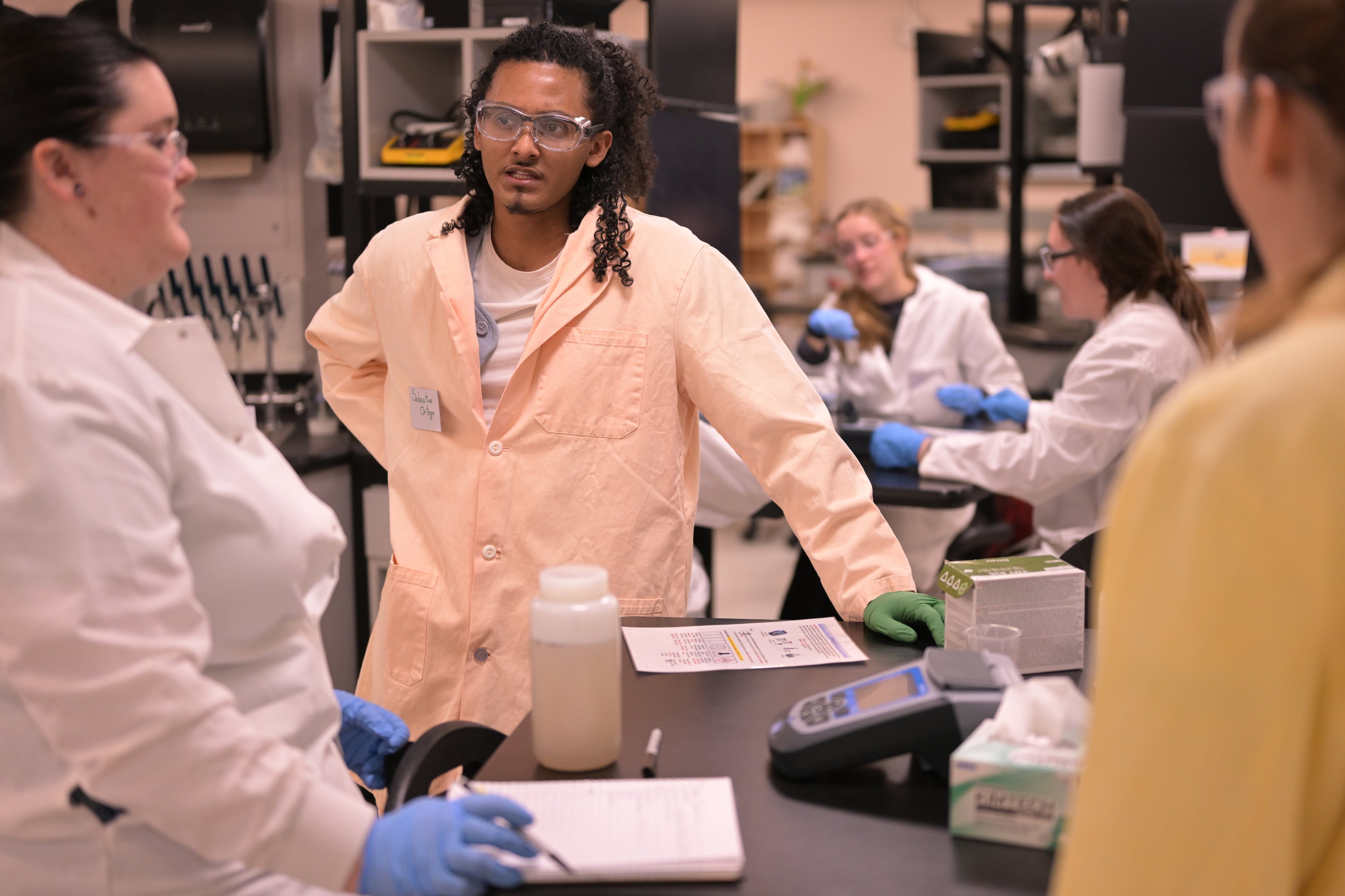 Students wearing lab coats and safety equipment work in a college laboratory.