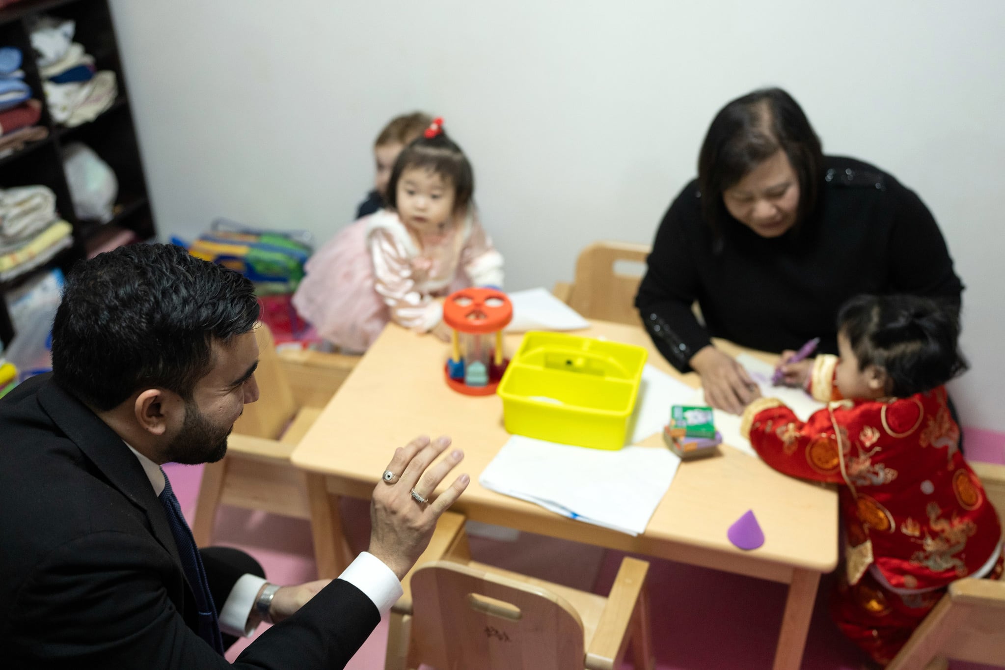 A photograph of a man in a suit kneeling next to a woman and three young children at a little table and chairs in a room.