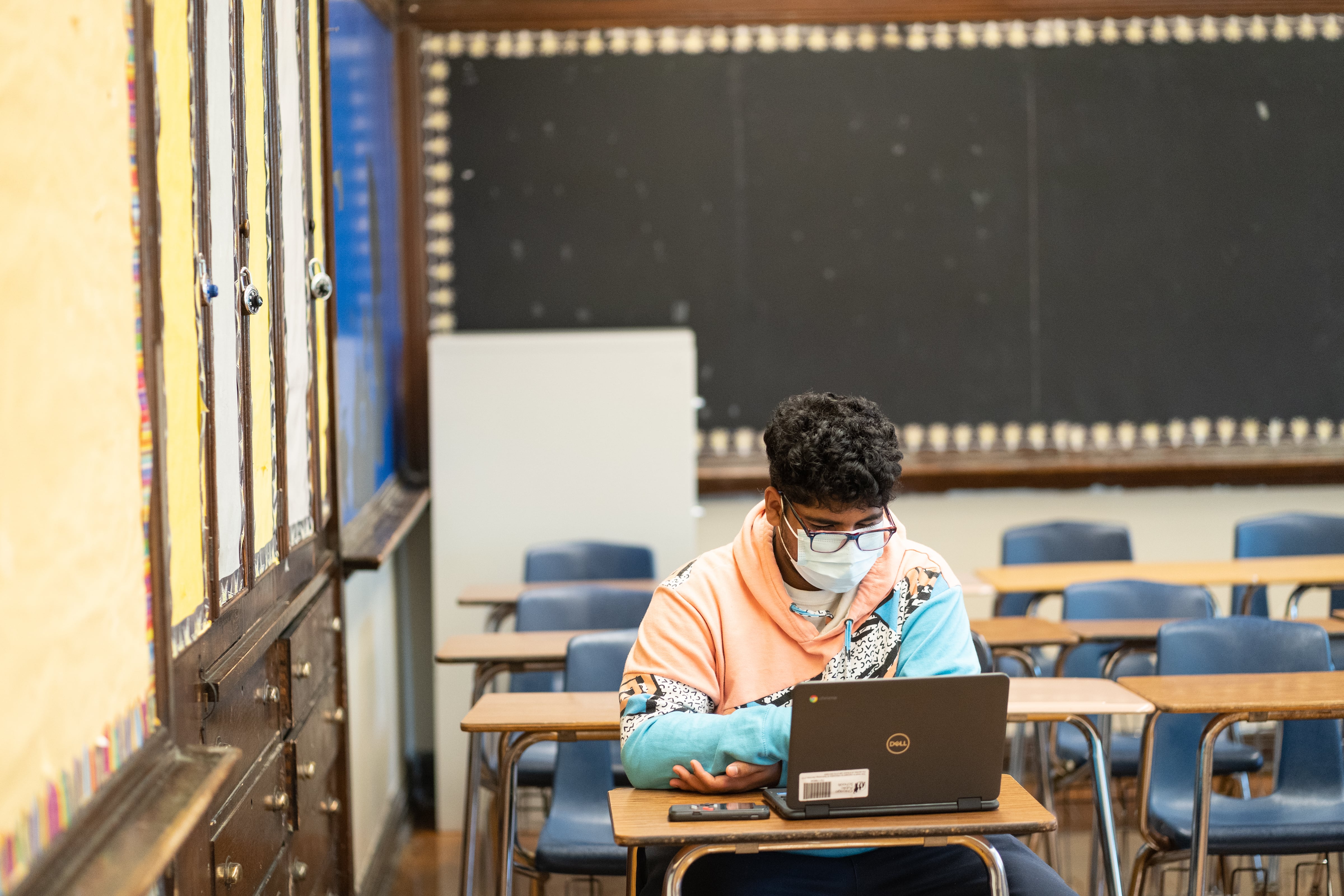 A young man wearing a pink, blue, and white hoodie works in an empty classroom on his computer.