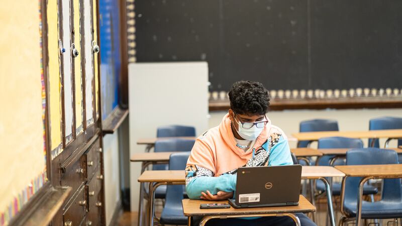 A young man wearing a pink, blue, and white hoodie works in an empty classroom on his computer.