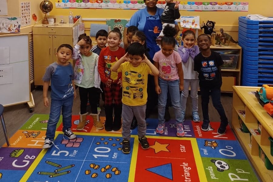 a teacher stands in a preschool classroom with roughly a dozen children