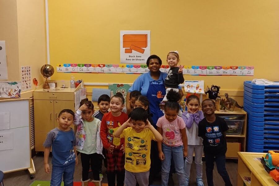 a teacher stands in a preschool classroom with roughly a dozen children