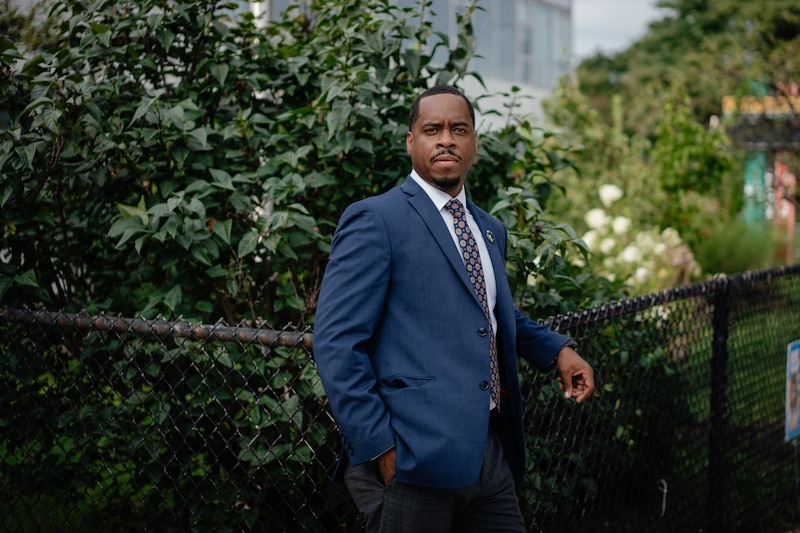 A photograph of a Black man in a blue suit posing for a photograph in front of a black chain linked fence with greenery and shrubs in the background.