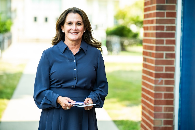 A woman wearing a dark dress and holding papers poses for a photograph outside near a red brick building.