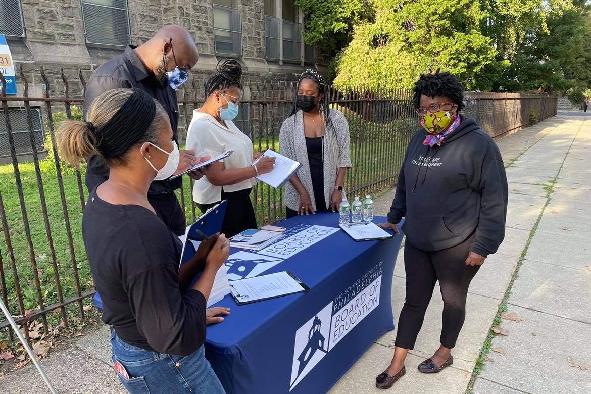 A group of volunteers stand around a table in front of a school with grass and bushes.