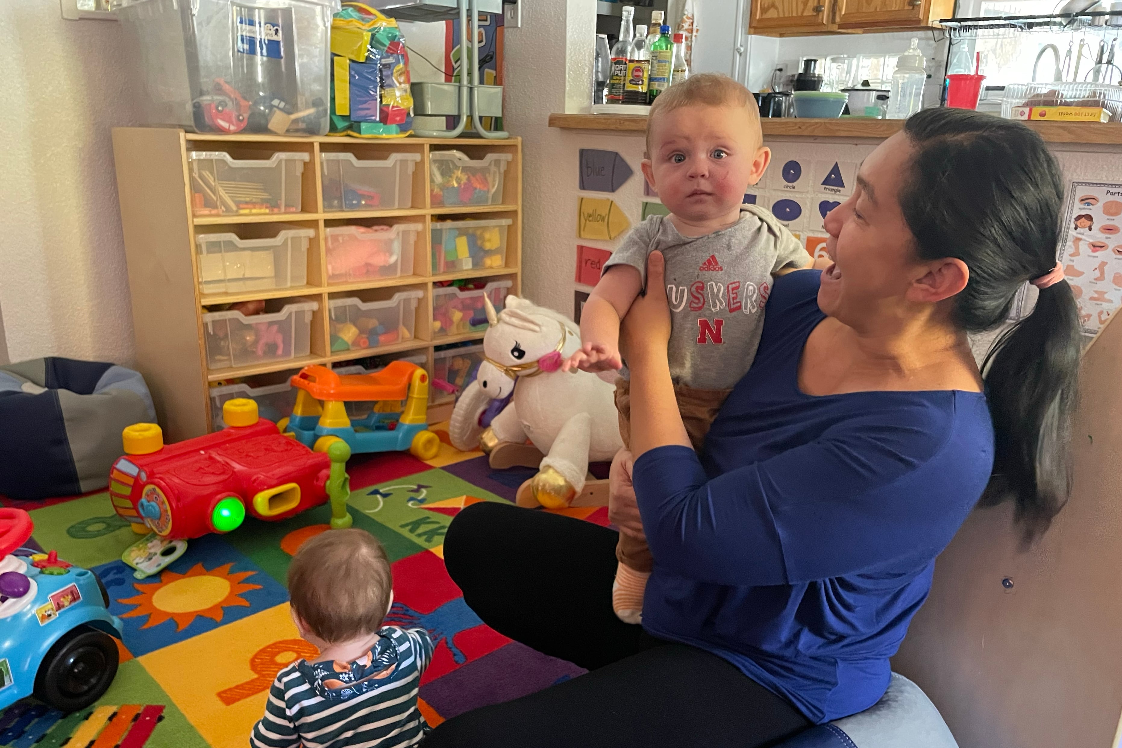 A woman with long dark hair and wearing a blue shirt holds a small child while another child sits at her feet.