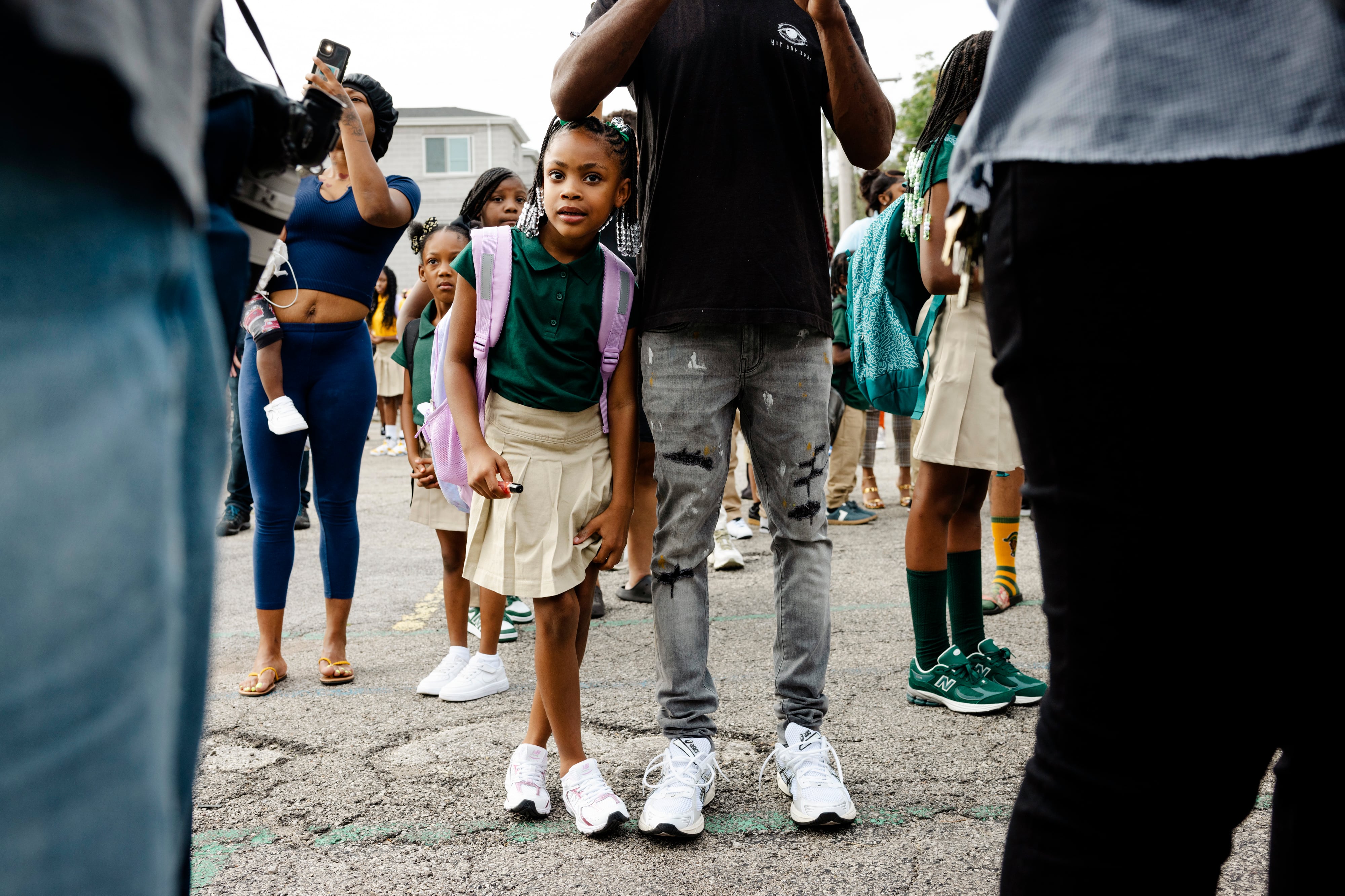 A photograph of a young Black student wearing a backpack and leaning against a person outside in a large crowd.