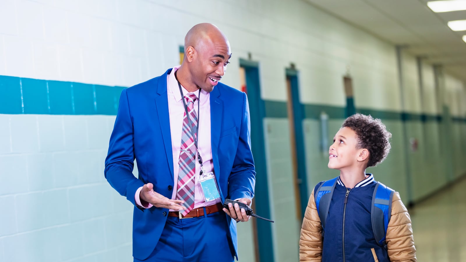 A man wearing a blue suit and a child wearing a blue and tan jacket walk thought a school hallway.