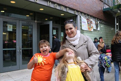 A woman wearing a grey jacket stands next to two young kids who are smiling and being silly.
