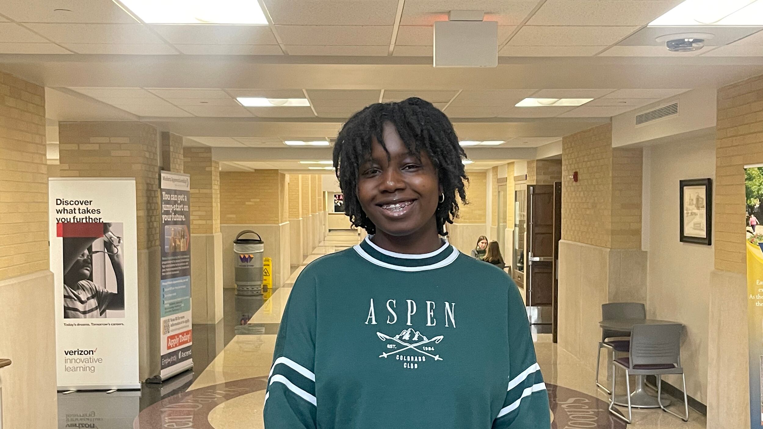 A young person stands in the center of a school hallway wearing a green sweater with the word "Aspen" on it.