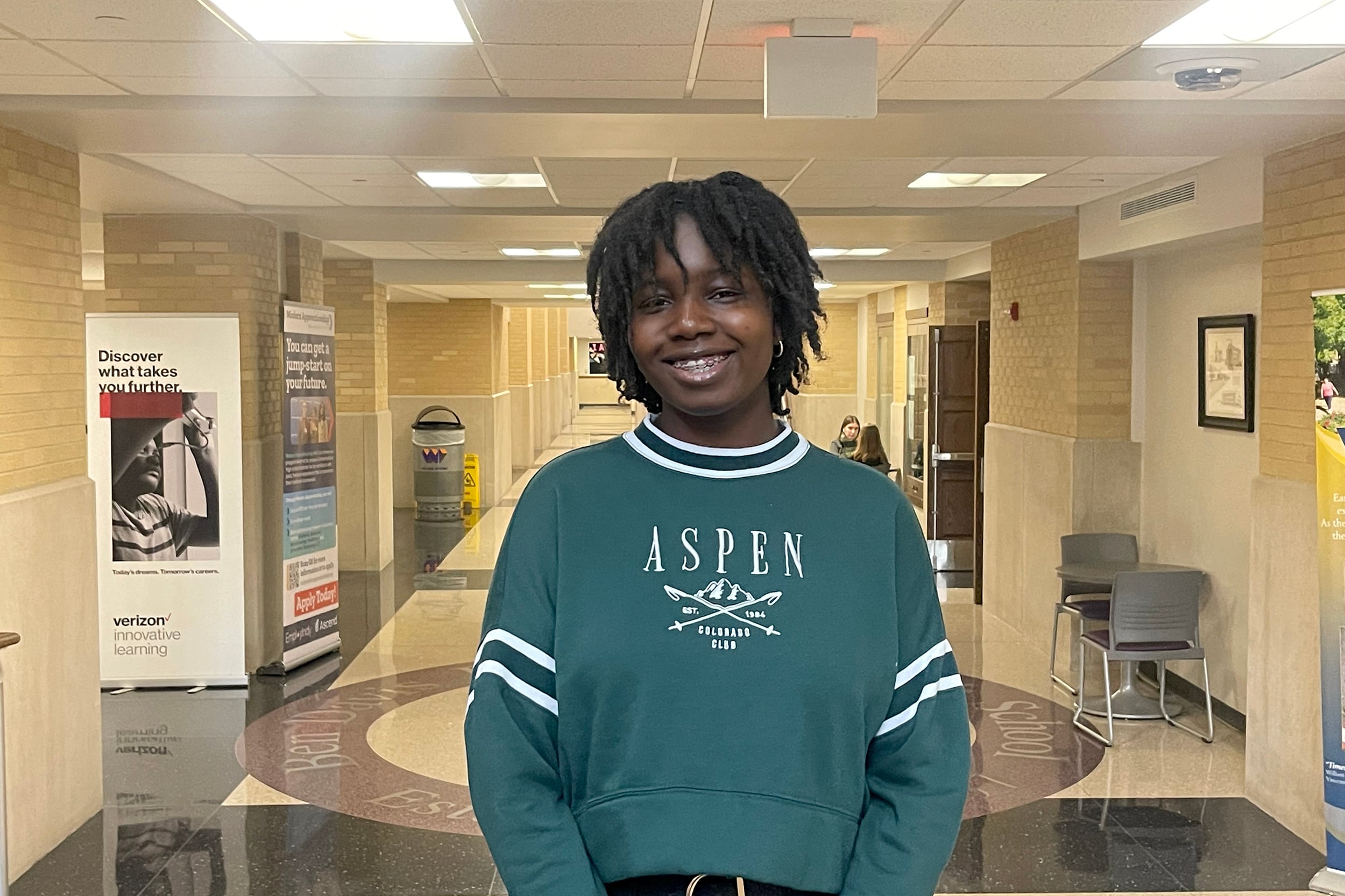 A young person stands in the center of a school hallway wearing a green sweater with the word "Aspen" on it.