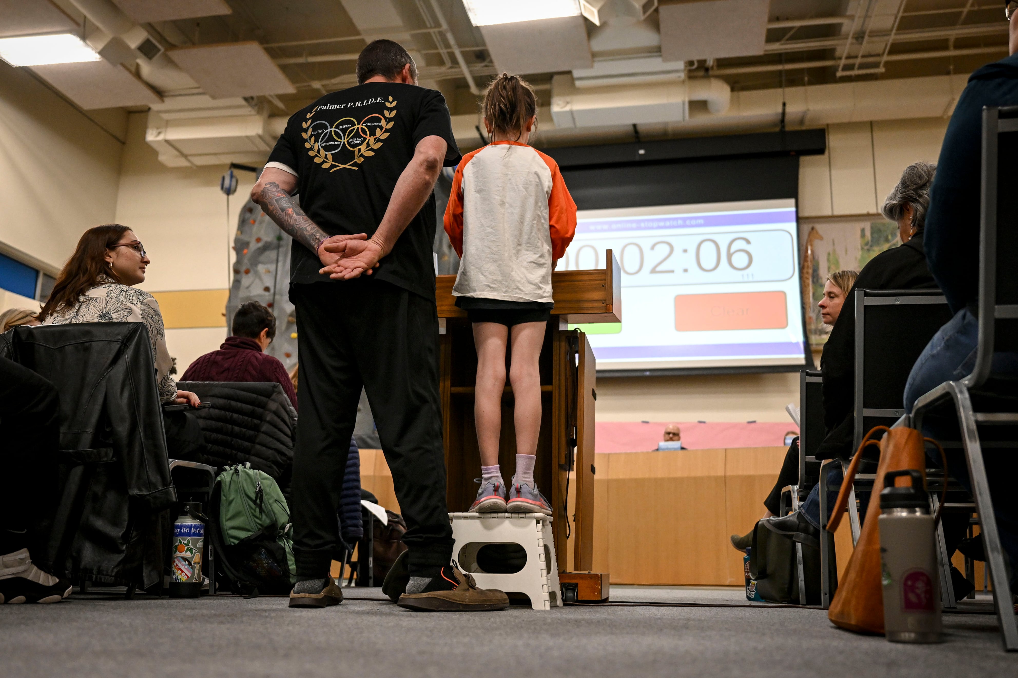 A young student stands on a step stool behind a podium and next to her dad with a projector screen in the background and people sitting in chairs on each side.