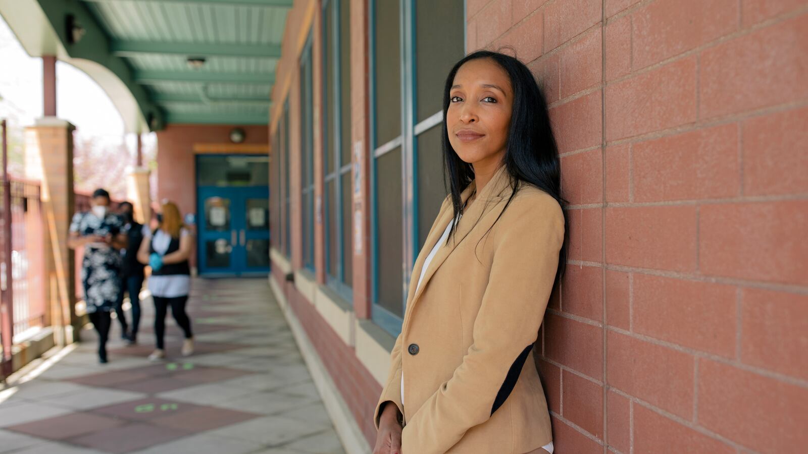 Yasirys Pichardo, wearing a tan pantsuit, stands against a red brick wall lined with windows while three women walk down the the path outside of P.S. 89.