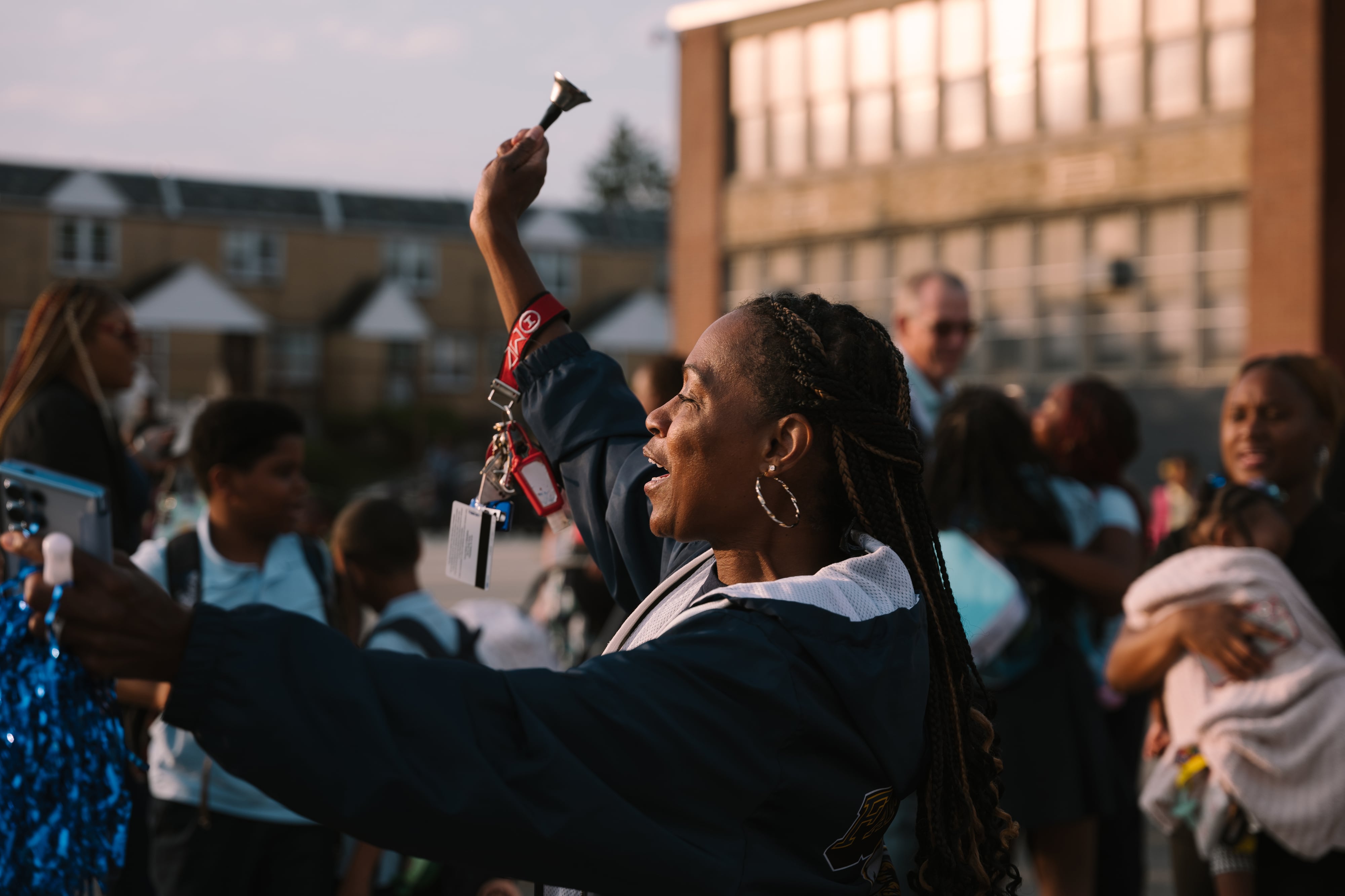 An adult rings a bell outside in a crowd of students and adults on the first day of school.