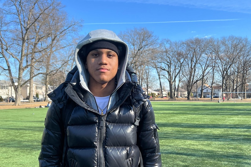 A high school student wearing a hoodie and a black jacket poses for a portrait outside on a cold, sunny day with a large green grassy area in the background.