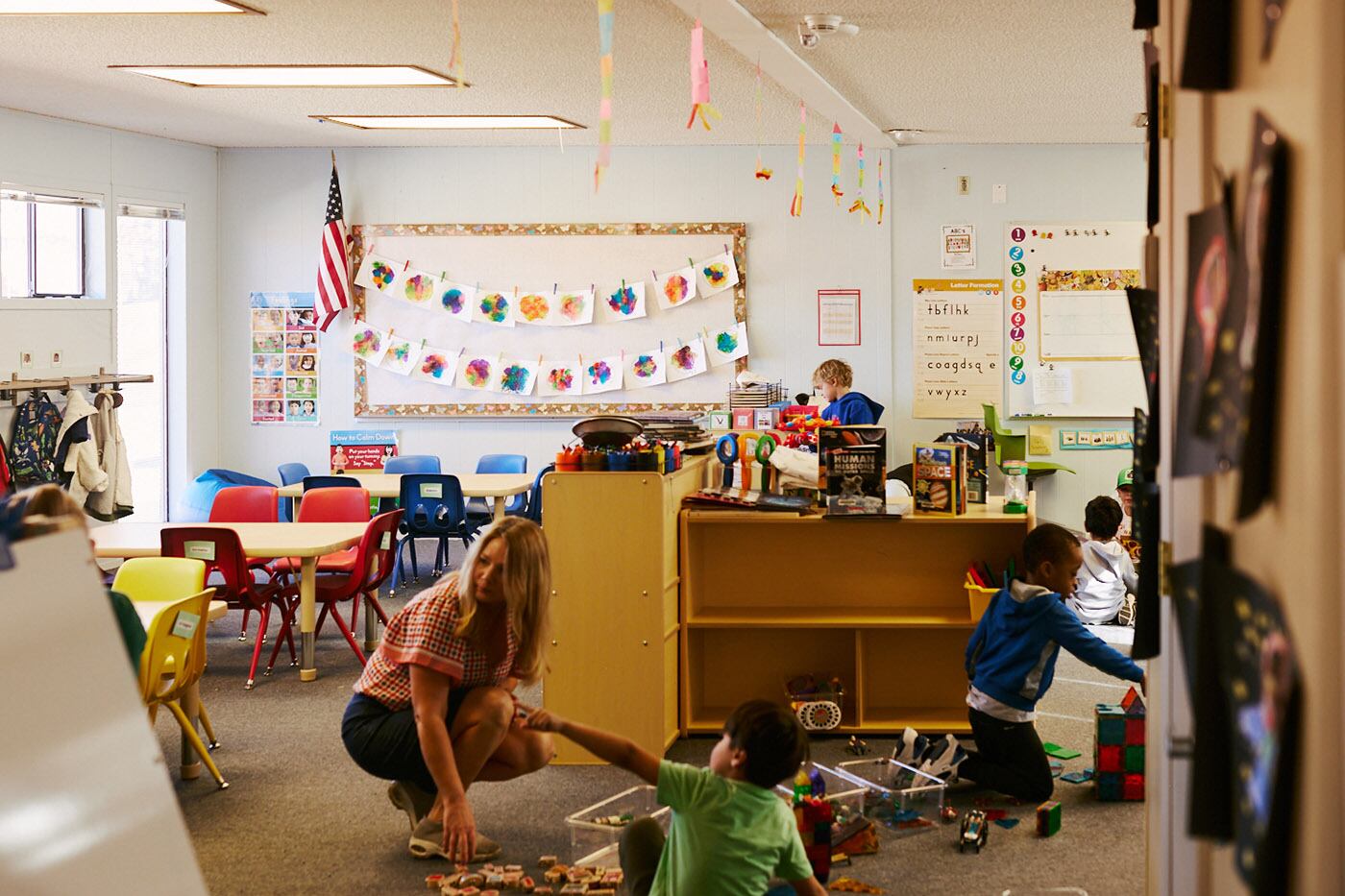 A teacher and student play on the ground with a colorful classroom in the background.
