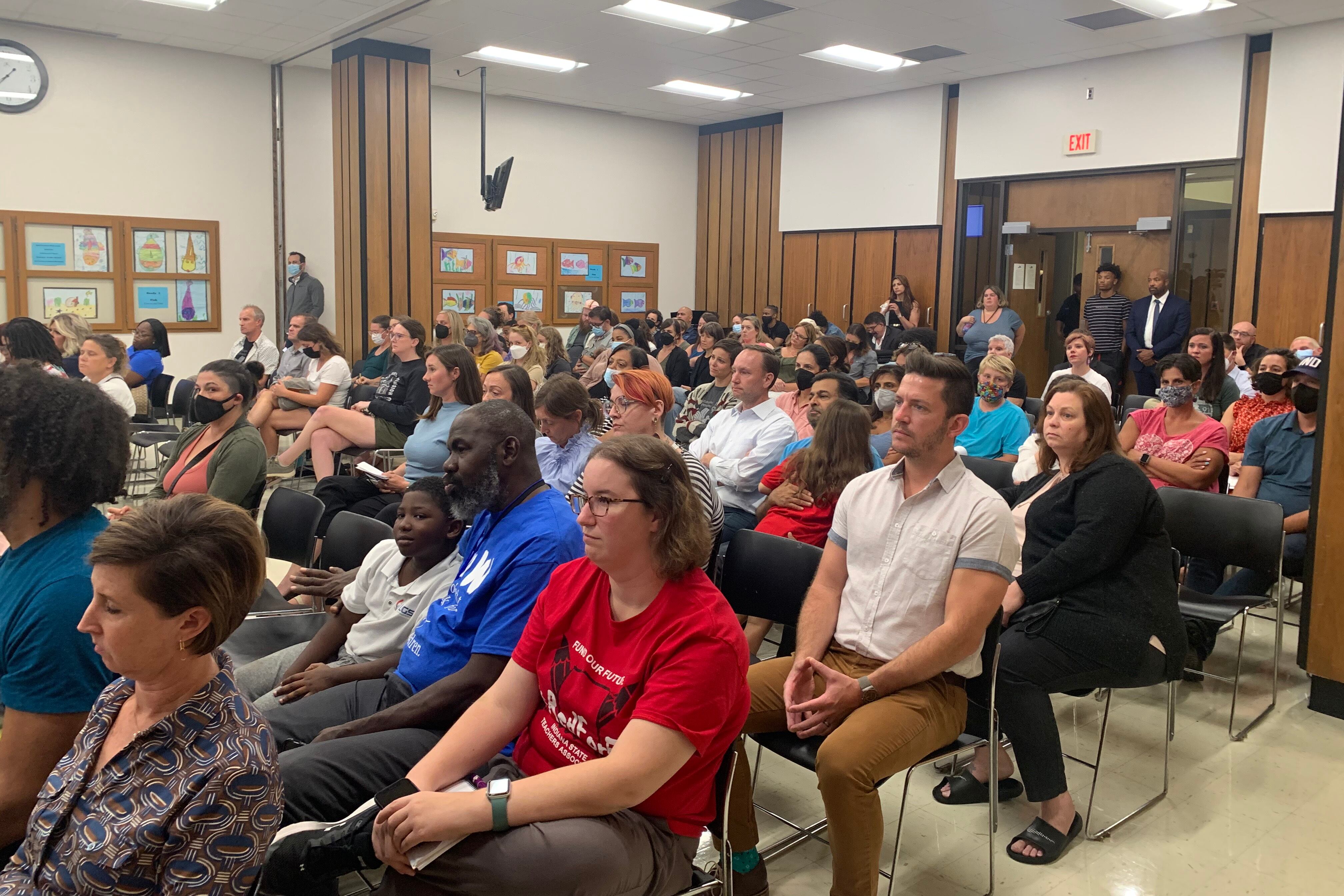 Dozens of adults sit in rows of chairs, watching a public meeting and facing in one direction.