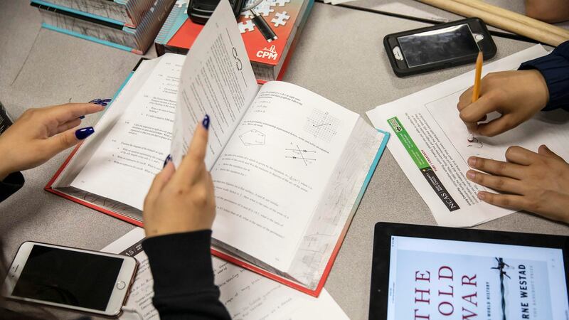 Hands of a student with black fingernail polish turn the pages of a math book, with an iPhone nearby. At the same table, the hands of another student erase a mark on a worksheet, with a tablet device depicting a book or lesson, “The Cold War,” nearby.