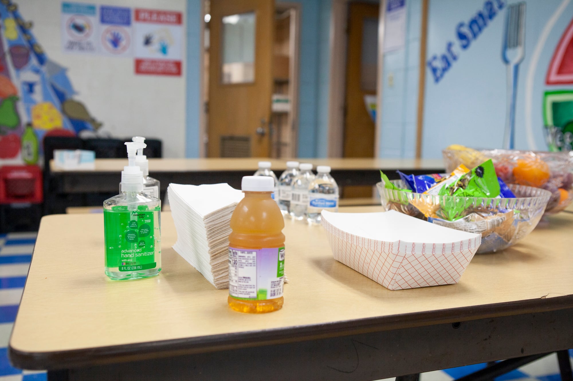 A bottle of hand sanitizer, a stack of napkins, apple juice, water bottles, and snacks on a table.
