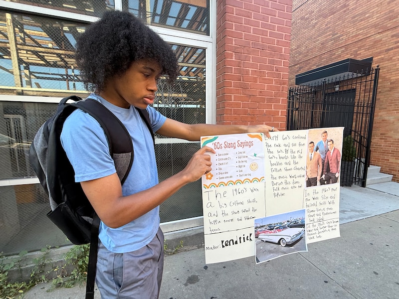 A photograph of a Black high school student with dark curly hair and wearing a blue shirt and a backpack holding up a white poster board with writing and photos on it.