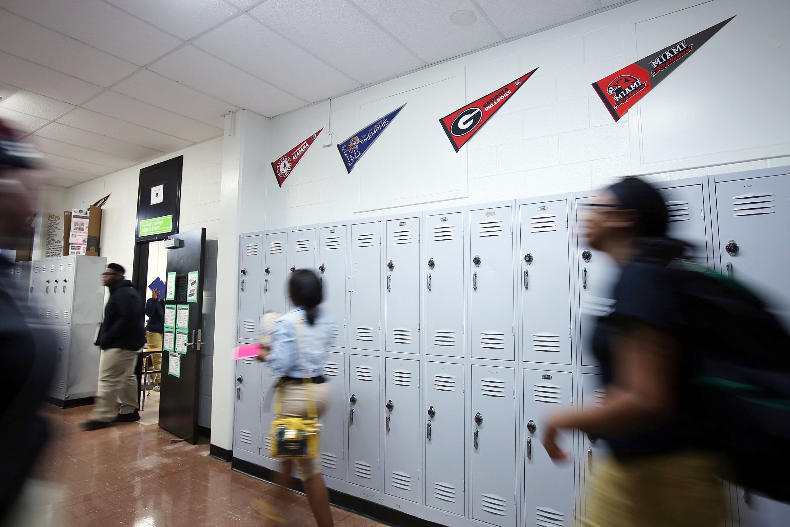 Students walk down a school hallway with lockers and college pendents on the wall.