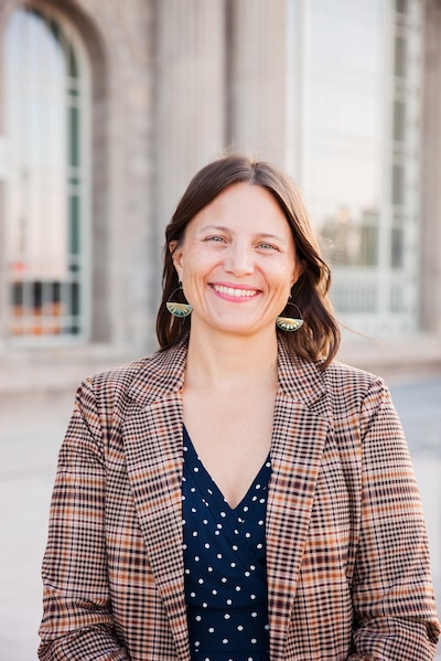 A white woman with dark hair and wearing a plaid suit jacket poses for a portrait in front of a large, stone building.